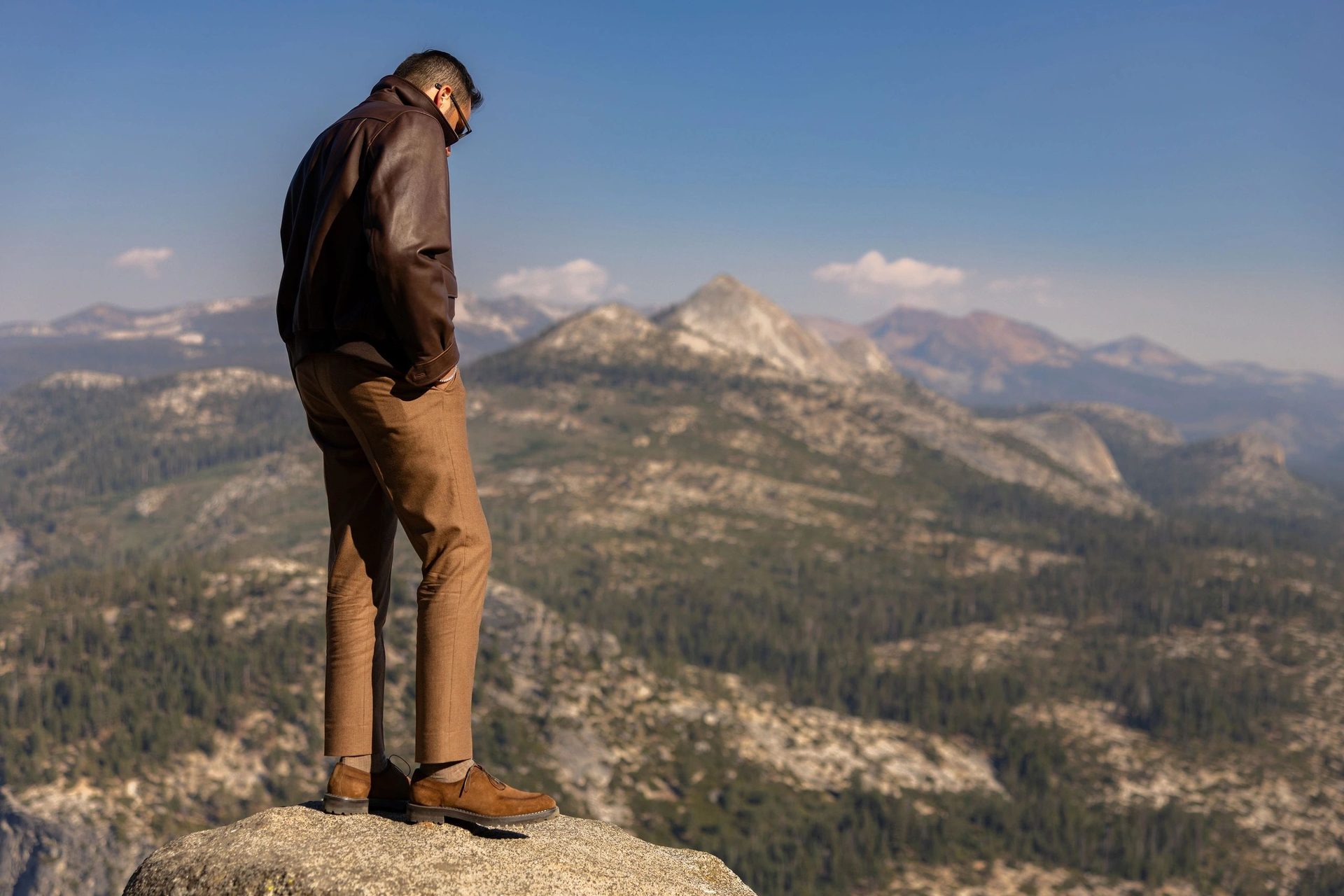 Model seen from behind in a brown leather jacket, tan trousers and suede shoes, standing on a granite ledge and looking down over forested ridges toward distant peaks