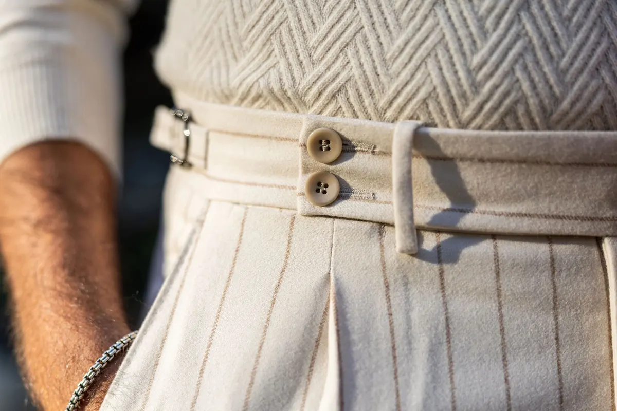 Close-up of cream pinstriped trousers with a double-button waistband and side adjusters, worn with a herringbone knit and a beaded bracelet