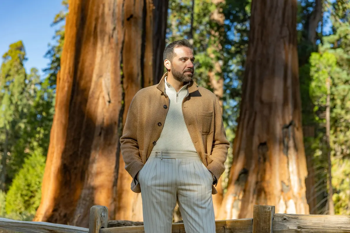 Model in a camel wool overshirt, cream half-zip knit and pinstriped trousers standing at a split-rail fence between two giant sequoias