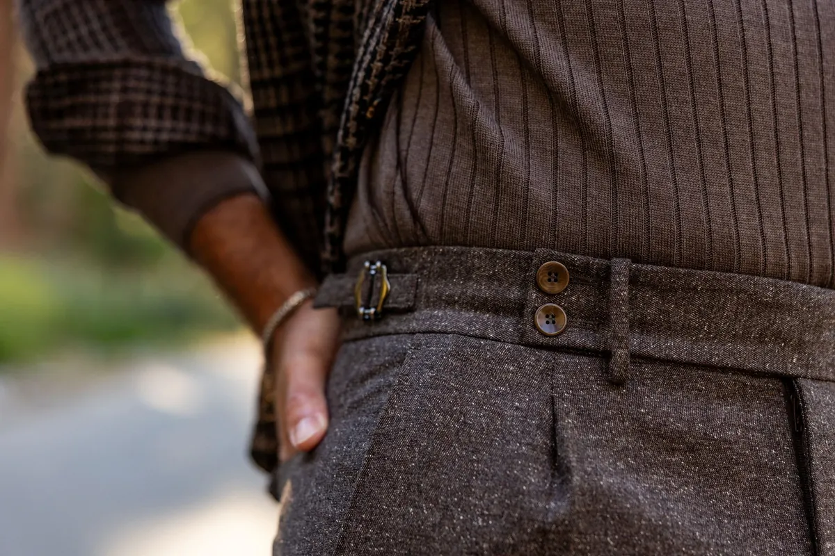 Close-up of a hand in the pocket of brown wool trousers with a buckled side adjuster, worn with a check overshirt and ribbed knit