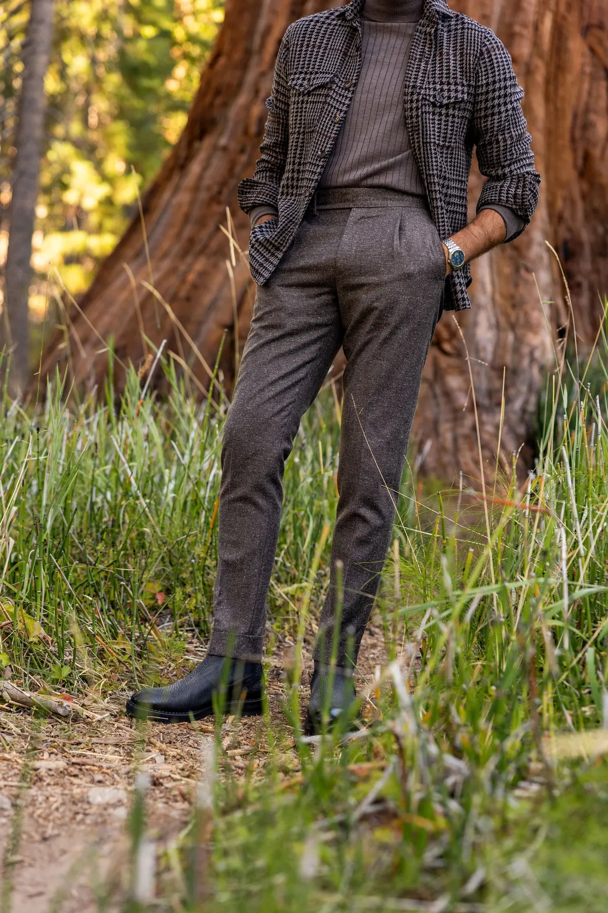 Model in a brown check overshirt and brown trousers standing in tall grass in front of a sequoia trunk