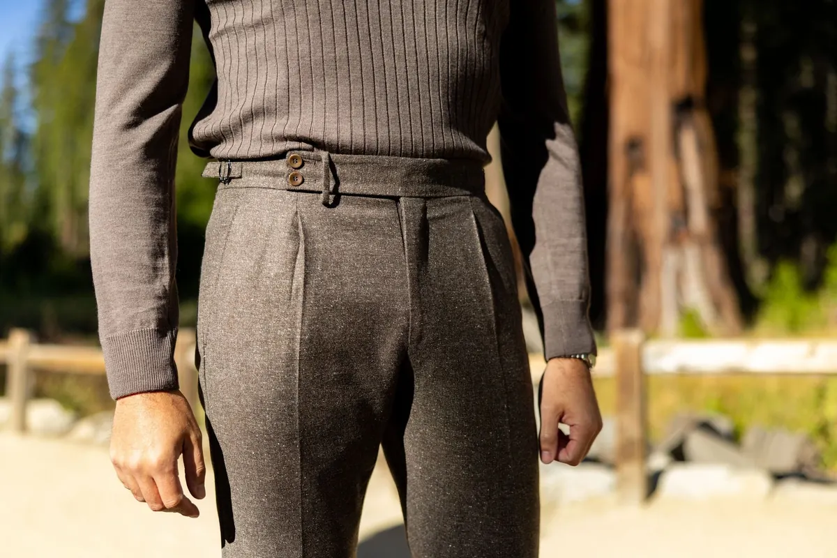 Brown ribbed knit and taupe flannel trousers with side adjusters, standing in front of a sequoia trunk and a split-rail fence