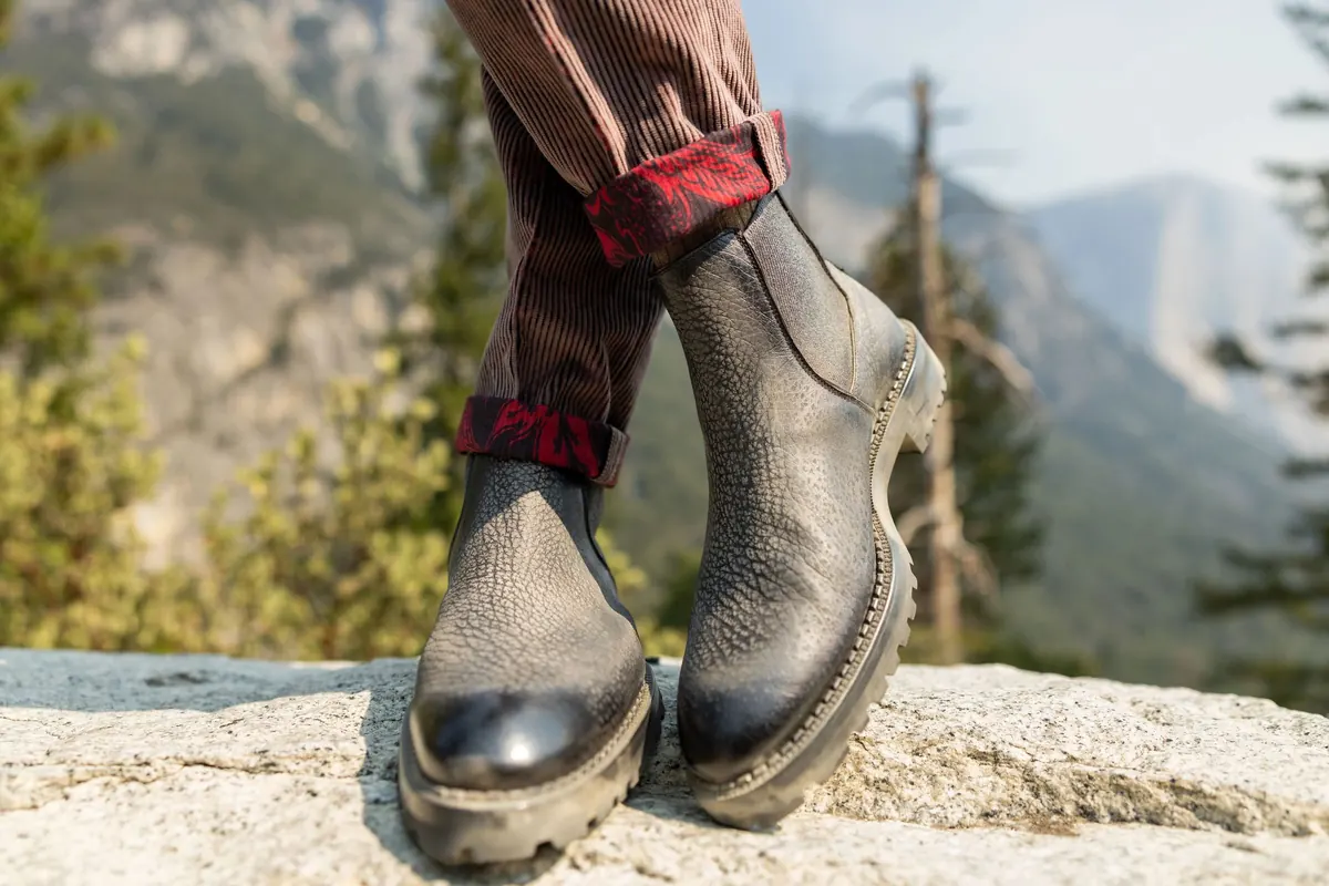 Close-up of grey pebbled-leather Chelsea boots with lug soles and corduroy trousers with red plaid turn-ups, posed on granite above a forested valley