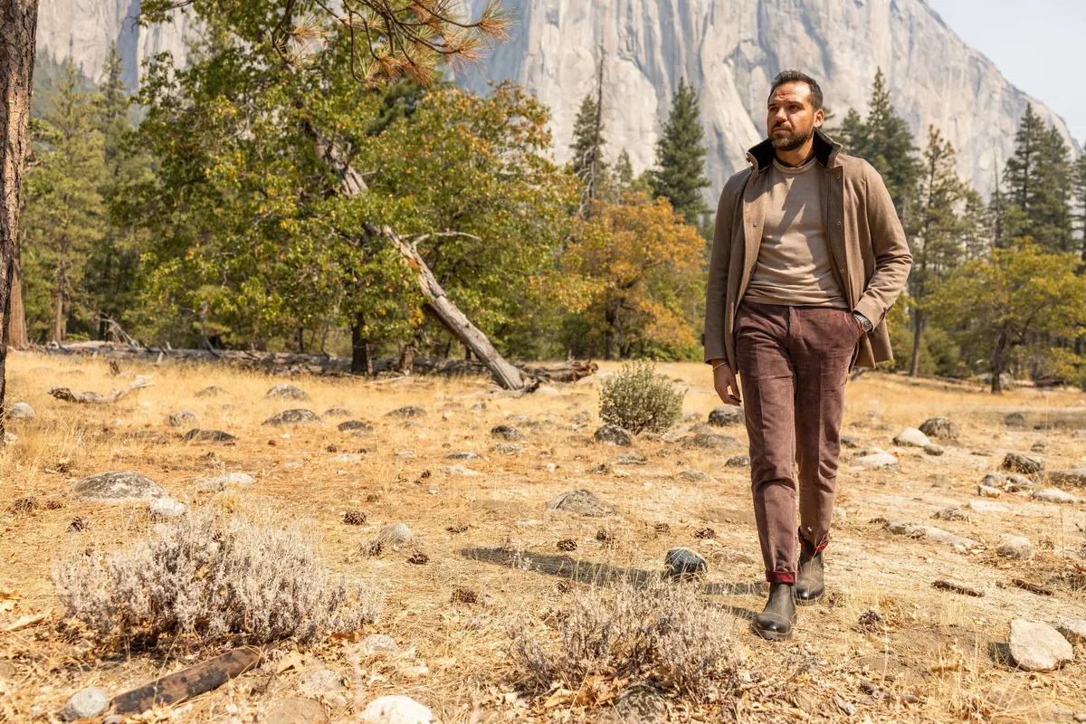 Model in a brown jacket and burgundy trousers walking through a sunlit meadow among pines and sagebrush, El Capitan behind