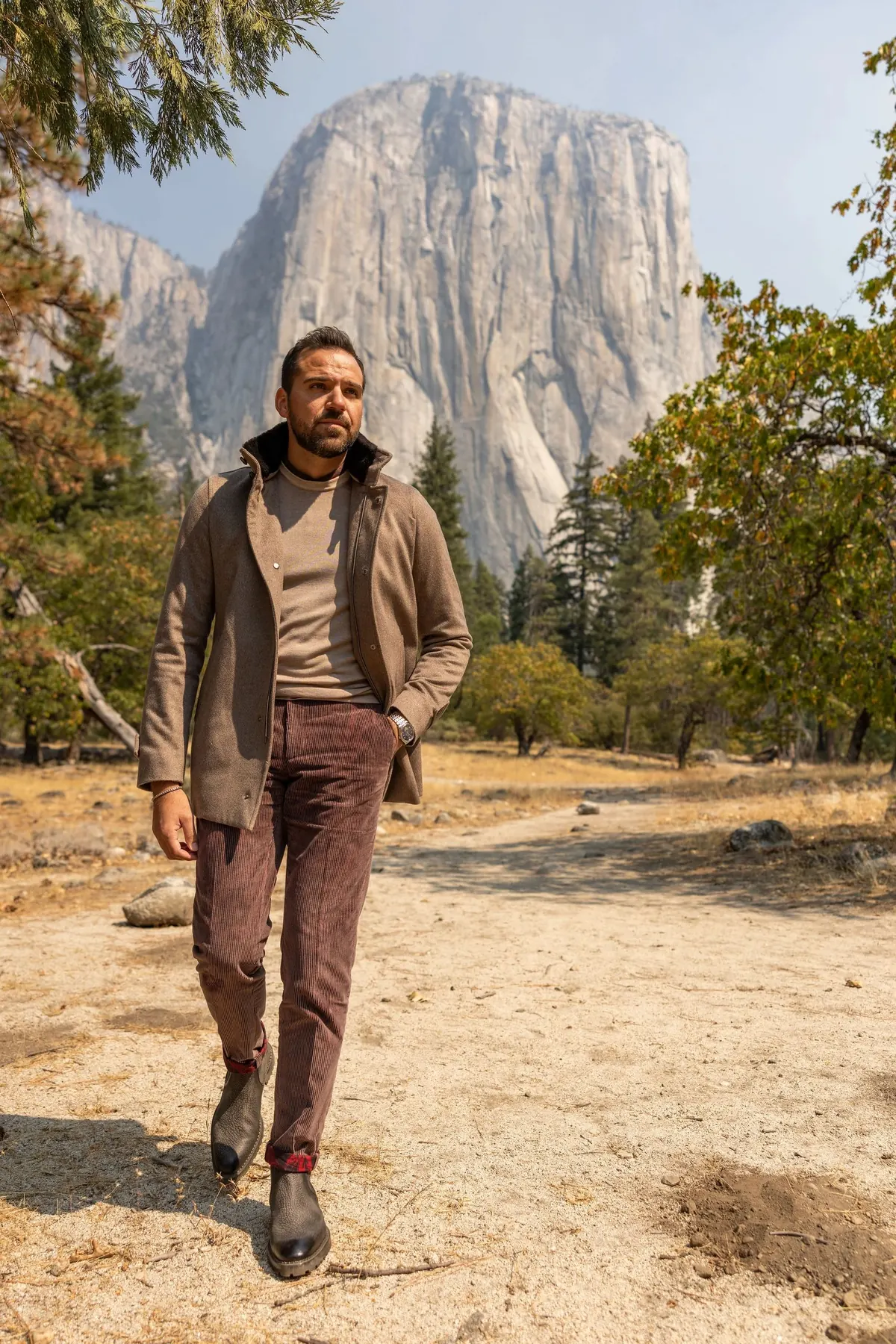 Model in a brown jacket over a knit sweater and burgundy trousers walking across a dry meadow toward the camera, El Capitan rising behind