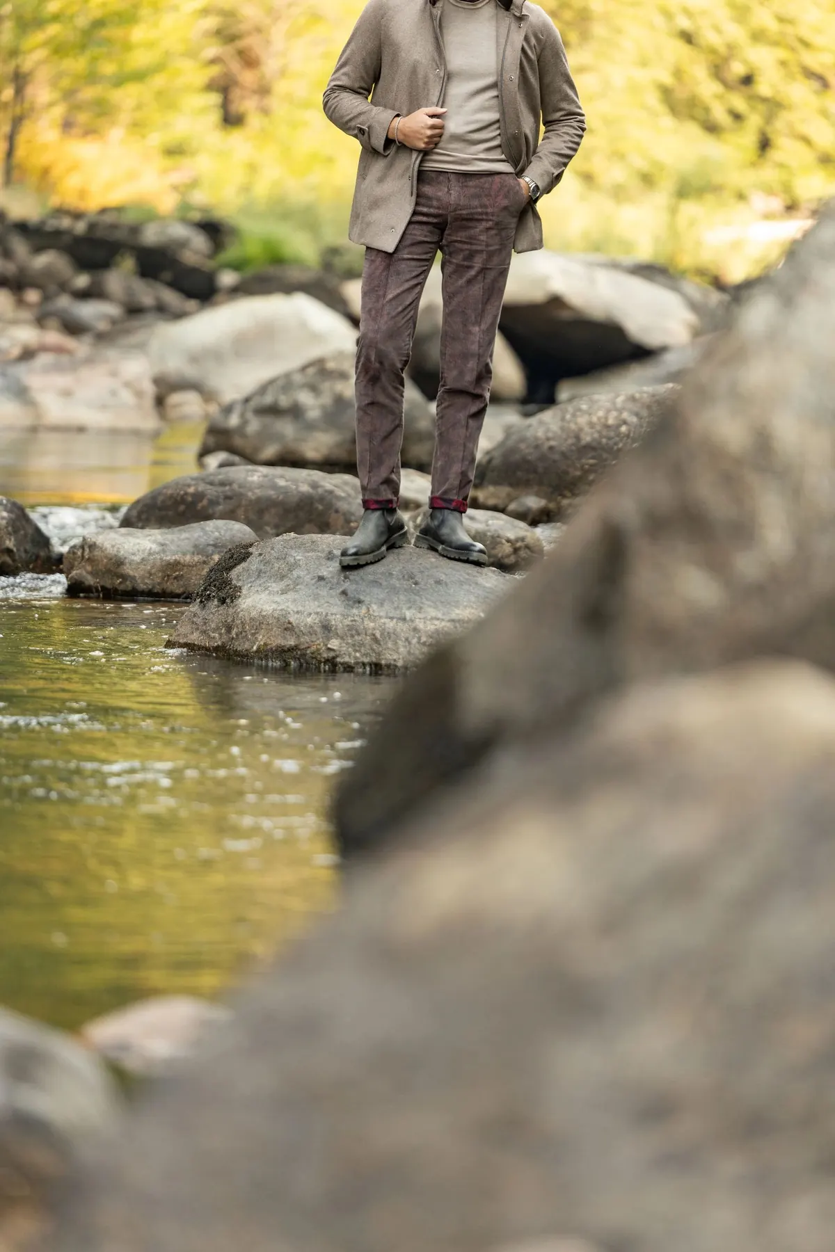 Model in a taupe jacket and burgundy trousers with plaid turn-ups standing on boulders above a clear river, a blurred rock in the foreground