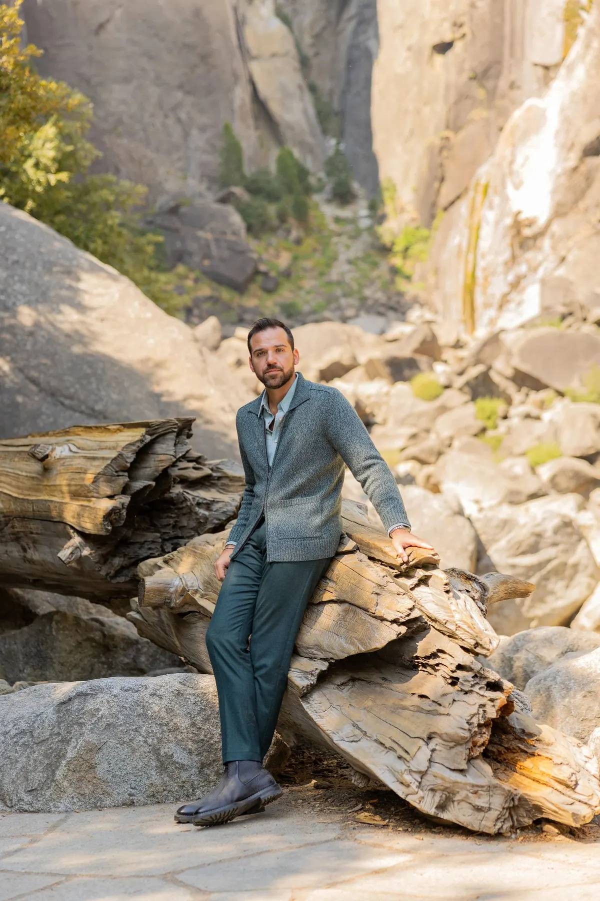 Model in a grey-green jacket and teal trousers seated on granite boulders and a weathered log, with rock walls behind