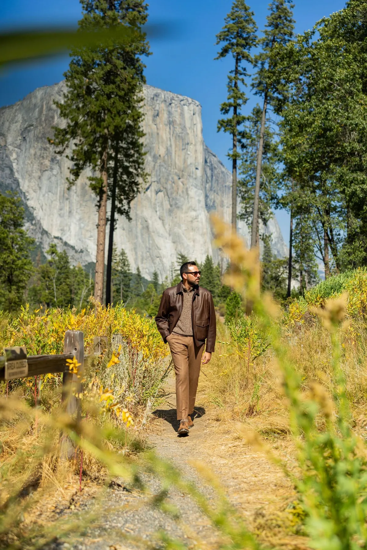 Model in a brown leather jacket, sunglasses and camel trousers walking a dirt trail through golden grasses, with El Capitan and tall pines behind under a blue sky