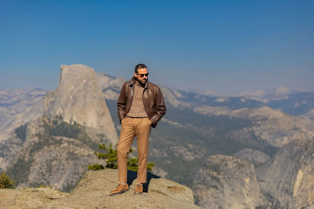 Model in a brown leather jacket, knit sweater and camel trousers standing on a granite ledge, with Half Dome and the high country behind