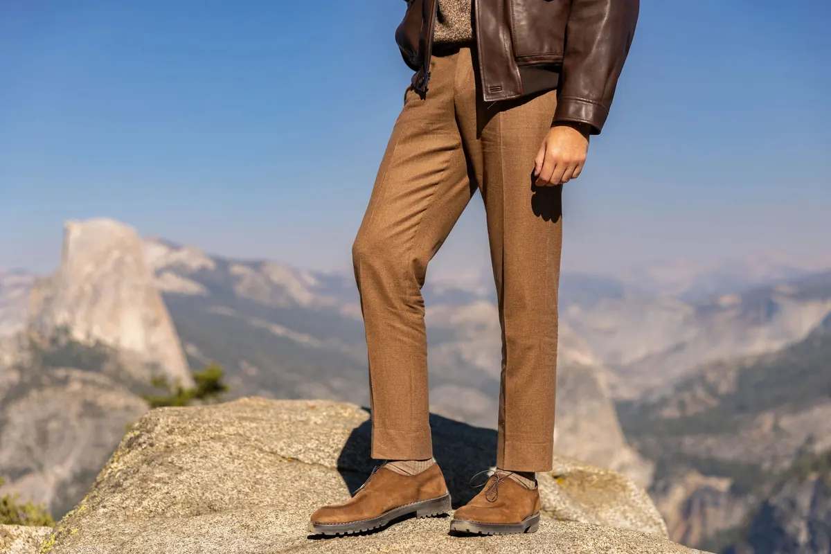 Camel trousers and brown suede shoes on a granite ledge below a brown leather jacket, with Half Dome in the distance