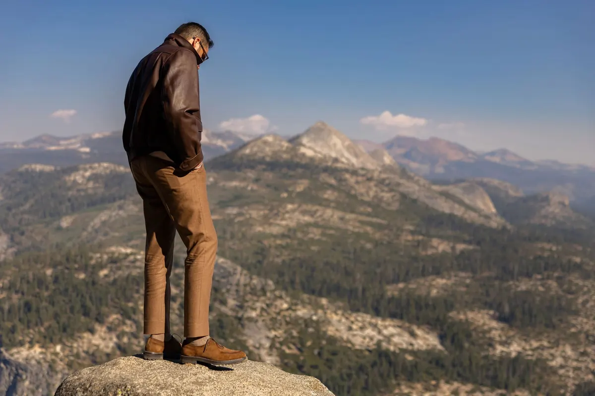 Model in a brown leather jacket and camel trousers on a rock outcrop, looking down over forested ridges toward distant peaks