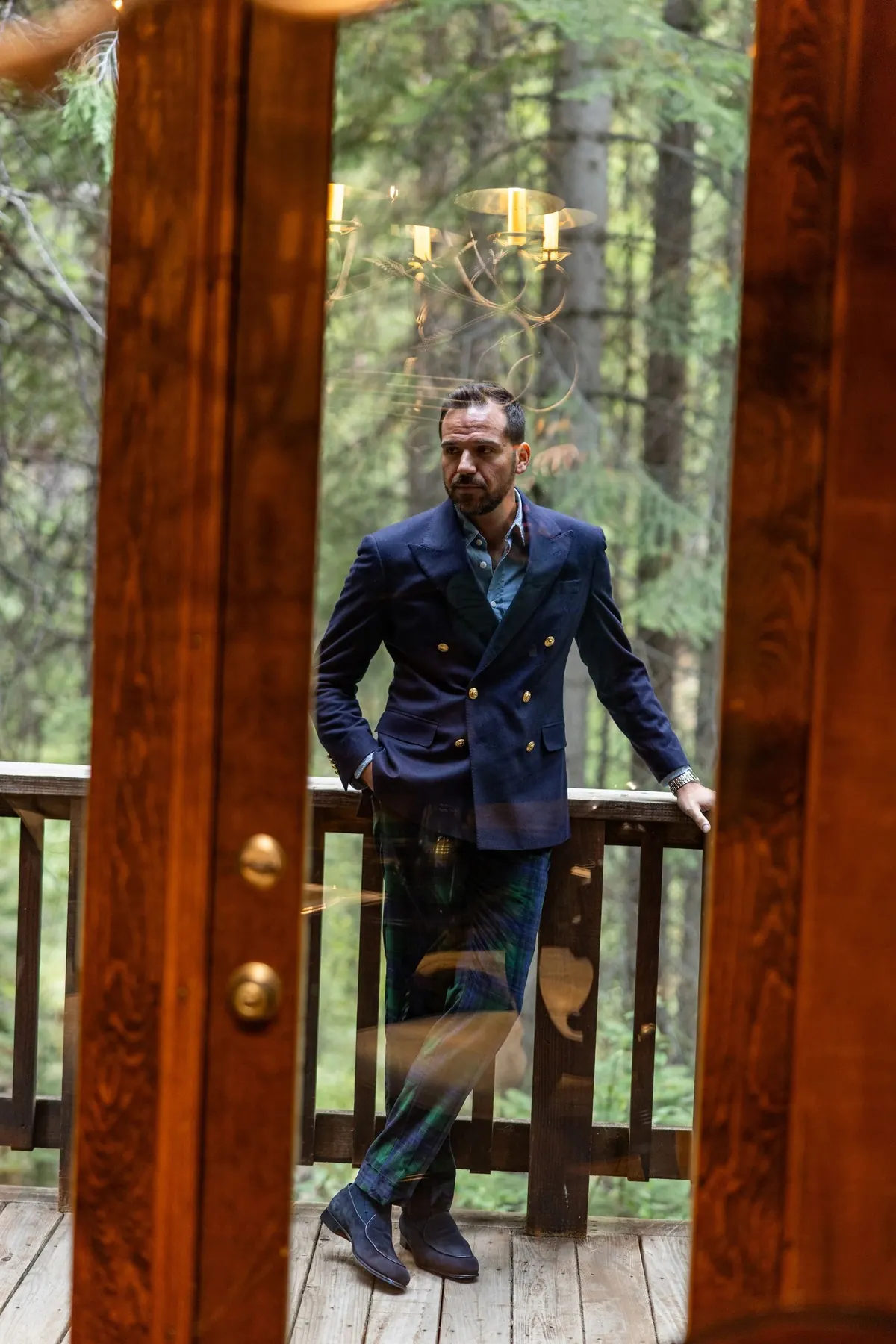 Model in a navy double-breasted blazer and tartan trousers framed by an open wooden door on a cabin deck among pines