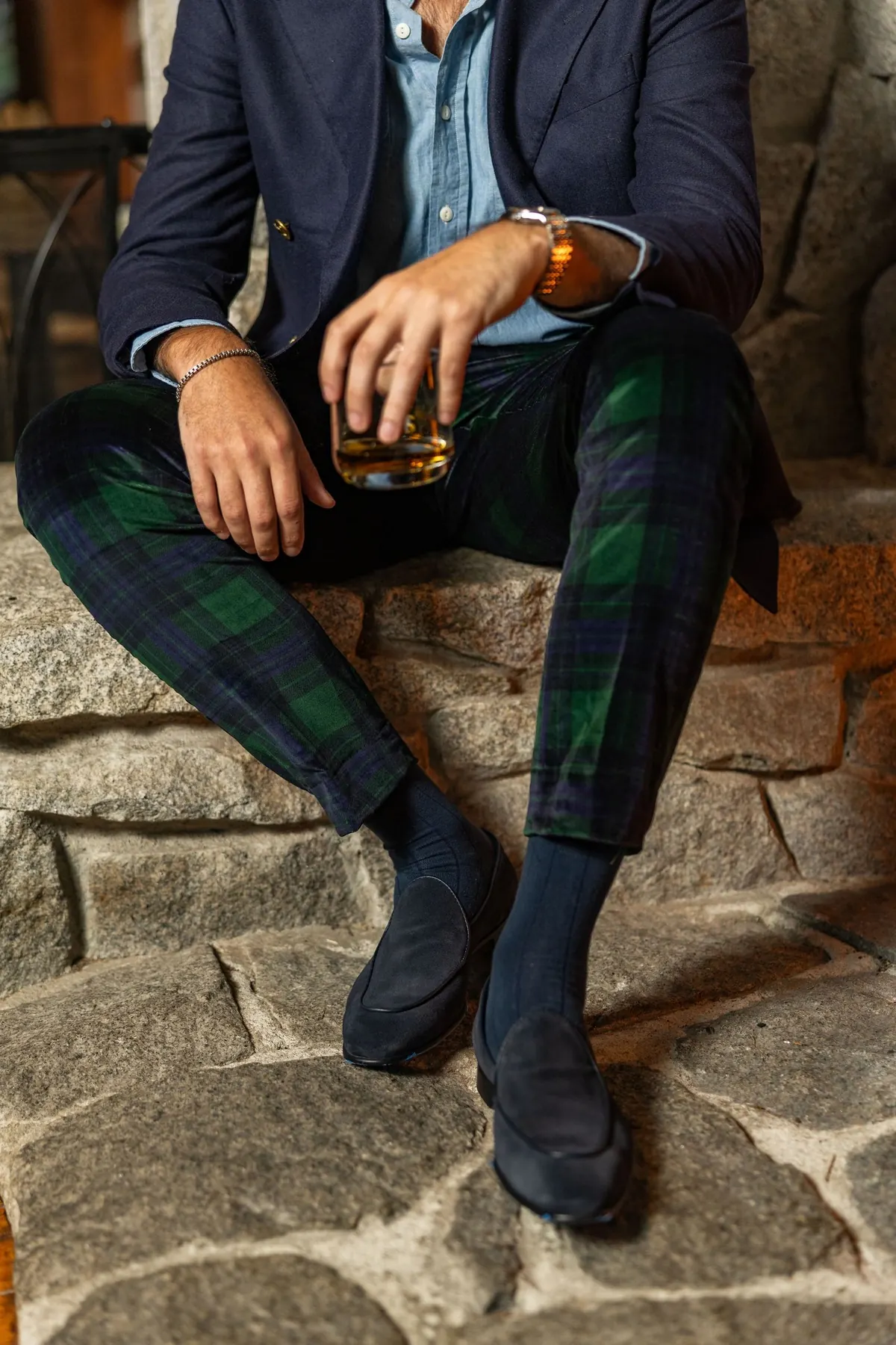 Seated on a stone ledge in a navy blazer and dark green tartan trousers, holding a glass of whiskey, wearing dark slippers