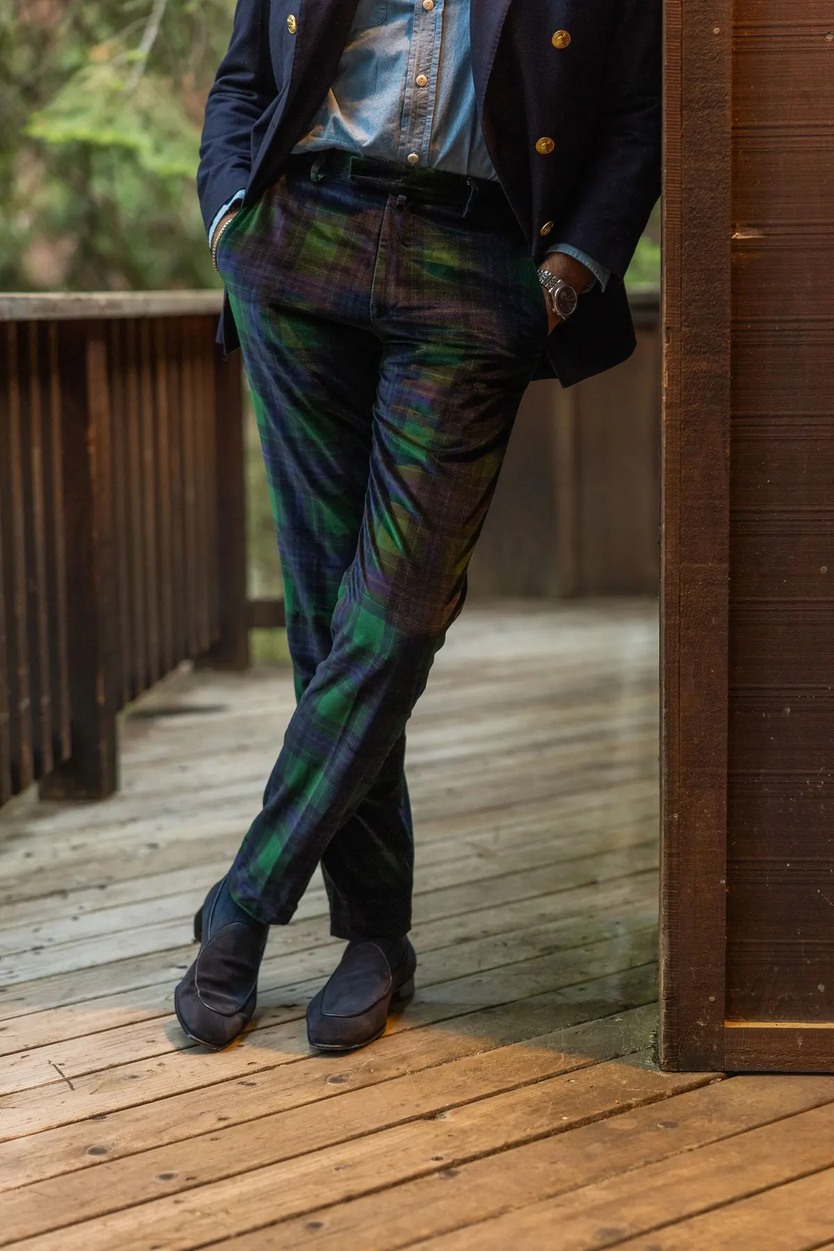 Navy blazer and blackwatch tartan trousers, legs crossed, leaning in a wooden doorway on a sunlit deck